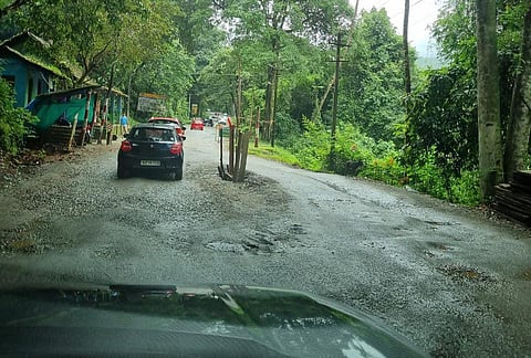 A narrow rural road with potholes, wet from rain, passes through a green forest area. Cars are moving along the uneven road, and small roadside huts with tarpaulin coverings are visible on the left. Trees and dense vegetation line both sides of the road.