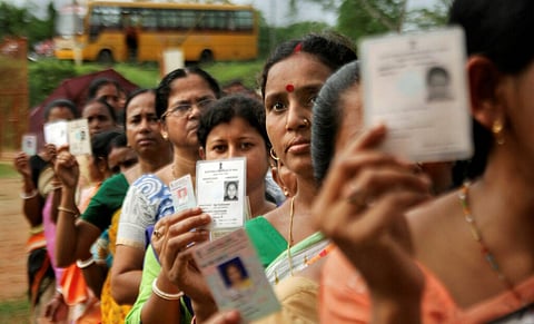 A medium-shot photo shows a long line of Indian women, many of them middle-aged, holding up their voter ID cards. The women are in a blurred outdoor setting with a yellow school bus visible in the background. They are wearing traditional Indian clothing in various colors and are waiting to cast their votes.
