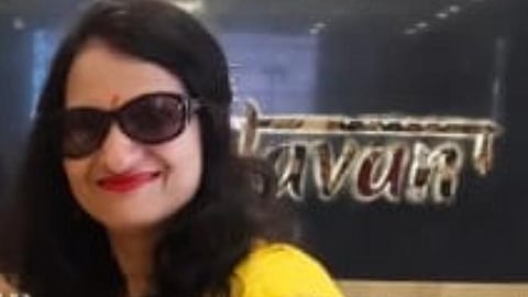 A woman wearing a bright yellow saree with a red embroidered blouse, accessorized with dark sunglasses and a small red bindi on her forehead. She has long black hair, styled loosely, and is smiling. She is holding a smartphone in her hand, with a mehndi design visible on her wrist and hand. The background shows part of a signboard and marble wall decor.
