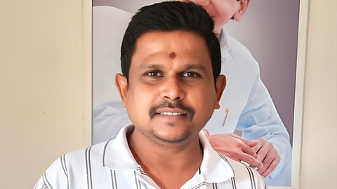 A man wearing a white shirt with black vertical stripes is standing indoors. He has short black hair, a mustache, and a small tilak on his forehead. Behind him, there is a large framed portrait of BRS president KCR in a white outfit, smiling with hands folded.
