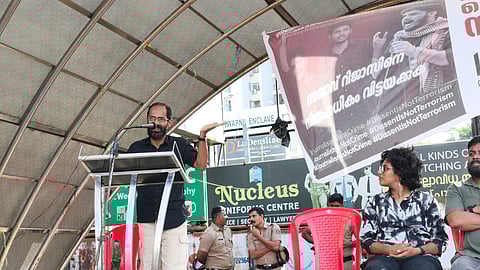 Siddique Kappan speaks from the podium placed on the left. He is wearing a black shirt and an off white pants