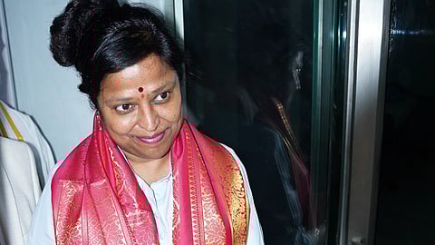 A woman wearing a white outfit and a bright pink silk shawl with gold patterns is cutting a green ribbon with scissors, marking the inauguration of an event or place. She has a red bindi on her forehead and is smiling slightly while looking at the ribbon.