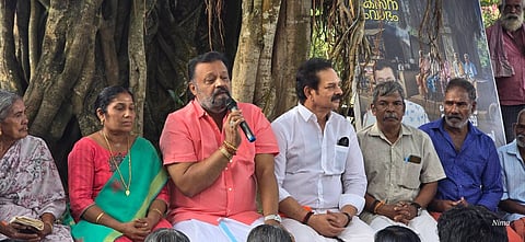 Suresh Gopi, Devan and others seated under a tree at a public outreach programme. Gopi is in a pink shirt and white mundu and is holding a mic