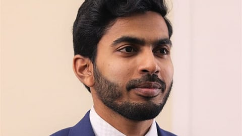 A man with short black hair and a trimmed beard is wearing a blue suit jacket, a white dress shirt, and a red patterned tie. He is looking slightly to the side with a composed expression, standing against a plain beige and white background.