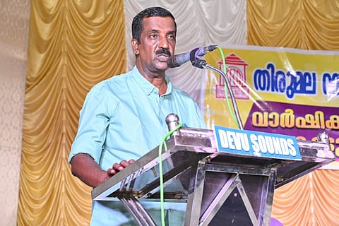 BJP councillor K Anilkumar, wearing a sky blue shirt, stands at a podium and speaks into the microphone.