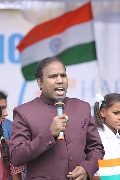 A man in a maroon Nehru jacket is speaking into a microphone while holding an Indian flag badge pinned to his chest. Behind him, the Indian national flag is prominently displayed. Children stand beside him, one wearing a white dress with a hair bow and another in a red school uniform. The scene appears to be part of an Independence Day or Republic Day celebration.