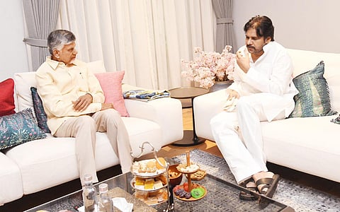 An image showing two prominent Indian political figures, N Chandrababu Naidu (left) and Pawan Kalyan (right), seated on a white sofa in a well-furnished room.

Naidu is wearing a light yellow shirt and beige trousers, sitting with his hands resting on his lap. Kalyan is dressed in a white kurta and pajamas, holding a small object (perhaps a medicine) to his mouth. A tiered platter with snacks, fruits, and dry fruits sits on a glass coffee table in front of them, along with some bottled water. The setting appears to be a formal meeting or visit.