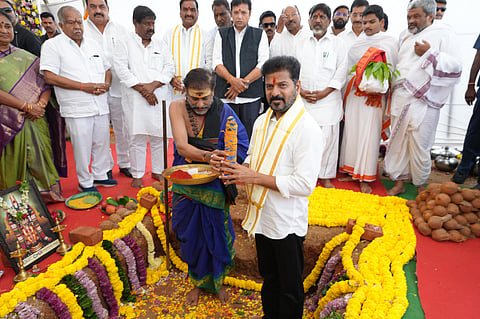 A photograph captures a traditional groundbreaking ceremony (Bhoomi Puja). The central figure, a man wearing a white shirt and a yellow and white scarf (Revanth Reddy, who is a prominent Indian politician), is standing in a partially dug pit adorned with yellow marigold garlands and surrounded by offerings like coconuts. He is looking down as a priest, who is kneeling and wearing traditional blue and saffron attire, is presenting a tray with colored powders (likely turmeric and kumkum) to him.

Behind them, a group of about a dozen men stand, mostly dressed in white traditional Indian attire (like dhoti and kurta), observing the ritual. One woman in a green saree is visible on the far left. The ground is covered with a red carpet. The ceremony appears to be a foundation-laying event for a new project.