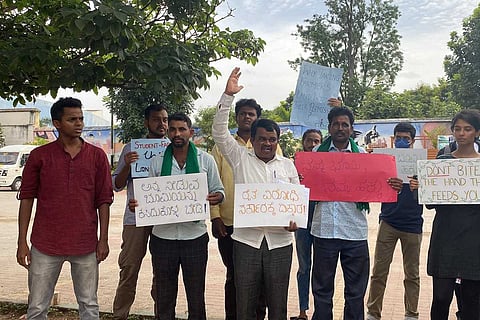 A group of farmers and supporters protest outdoors in Devanahalli, Karnataka. They hold placards in Kannada and English, with messages such as “Don’t bite the hand that feeds you” and “Why taking farmers’ land without their permission?” One protester raises his hand while others stand with serious expressions.