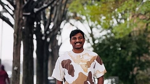 A full-body photo of a man standing on a snowy sidewalk, smiling directly at the camera. He is wearing a camo-patterned t-shirt with white, tan, and brown shapes, and black pants. His hands are in his pockets. The sidewalk is covered in a light layer of snow, and he is flanked by a line of tall, bare trees that form an avenue, with some green foliage visible in the background on the right. The overall setting suggests a winter day in a suburban or park-like area.