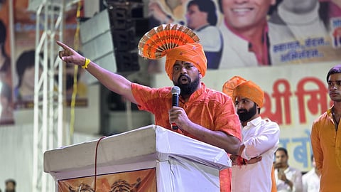 A man with a beard, dressed in an orange kurta and turban, stands at a podium, holding a microphone and pointing his right arm up and out to the left. His turban is decorated with a fan-like orange plume. A portrait of a man in an orange outfit, framed by images of tigers, is visible on the front of the podium. Two other men in white kurtas and orange turbans stand to his right. The background is blurred, showing a stage or banner with some text and faces.