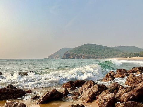 A high-angle photo shows a wave breaking on the shore of a beach, with foamy white water crashing over large, dark brown and reddish rocks in the foreground. The foreground also includes wet sand. Beyond the immediate wave, the Bay of Bengal stretches out under a hazy, pale blue sky. In the background, two large, green-covered hills dominate the scene, sloping down toward the sea and meeting the beach on the right side of the image. The sunlight is bright but diffuse.