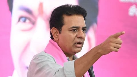 A close-up shot of K. T. Rama Rao (KTR), an Indian politician, wearing a light gray collared shirt and a pink scarf/kanduva, holding a microphone and pointing his right hand forward and slightly up, with a serious expression while speaking at a political rally. A large, blurred pink backdrop features a grayscale image of an older man's face, partially visible behind him. Other people are seated or standing in the background, mostly blurred.
