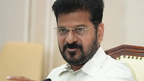 A head-and-shoulders, medium-close shot of Telangana Chief Minister Revanth Reddy, seated at a desk or table, speaking or making a point.

He is wearing a light-colored, possibly off-white, collared shirt. He has a dark beard and mustache, dark hair, and is wearing clear-framed rectangular glasses. He is looking slightly off-camera to the right with a serious expression and is extending his right arm forward, pointing with his index finger. A black microphone is visible just in front of him. The background is indoors and includes blurred details of light-colored furniture and a wall. A water bottle is visible on the right side of the desk.