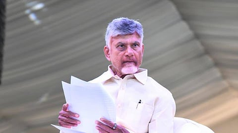 A low-angle, full-body shot of an older man with gray hair, N. Chandrababu Naidu, seated in a large, plush, off-white armchair. He is wearing an off-white button-down shirt and matching trousers. He is holding a stack of white papers in his hands and looking straight ahead with a focused expression. He is wearing black slip-on dress shoes. The background is a brightly lit, slightly out-of-focus indoor setting.