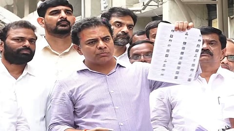 A medium close-up photo shows K. T. Rama Rao (KTR), dressed in a striped light purple shirt, standing amongst a group of men, all of whom appear to be politicians or party workers.

KTR is holding up a sheet of voter rolls, which he is presenting to the camera. His expression is serious. The other men are dressed in white shirts and look on intently. The background is an out-of-focus outdoor scene, possibly near a building.