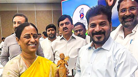 
A full-length photo showing Konda Surekha, a woman in a bright yellow saree with a golden border and a patterned blouse, presenting a wooden figurine of a woman holding a bowl to Ponguleti Revanth Reddy, a man wearing a light blue collared shirt and light gray trousers.

Both Surekha and Revanth Reddy are smiling at the camera. They are standing indoors on a green carpet at what appears to be an official event. Several other men in formal or semi-formal attire are visible in the background, with one man's face partially cropped on the right. A banner with a blue and white design and the number "1" is visible behind them.