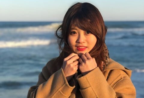 Baek Se-hee standing by the seaside, wearing a brown coat and smiling softly at the camera with her hands near her face. The ocean and gentle waves form the background.