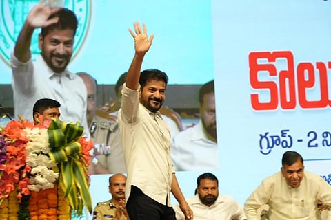 Telangana Chief Minister Revanth Reddy waving to the crowd while walking across a stage. He is wearing a white shirt and dark pants, has a beard, and is smiling. In the foreground to the left is a large floral bouquet. Other people are seated on the stage behind him, and to the right, a large Telugu sign is visible.