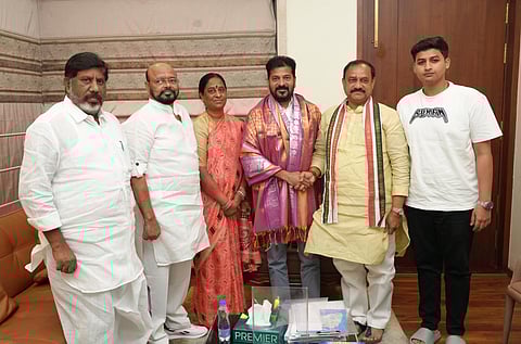 A formal group photograph indoors. Six people are standing together. Revanth Reddy, the Chief Minister of Telangana, is in the center, wearing a purple and pink patterned shawl. To his left is a woman in a peach saree and two men in white kurtas. To his right is a man in a yellow kurta and a young man in a white t-shirt.