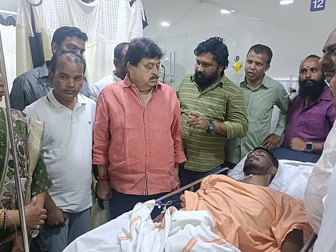 A group of men stand around a man lying on a hospital bed. The man on the bed is wearing an orange garment and has medical tubing attached to him. A prominent man in the center, wearing a salmon-colored shirt, is looking down at the injured man. Other men are gathered around them, some looking concerned. The setting appears to be a hospital room.

The image is a photo of BJP leader Ramchander Rao (in the salmon-colored shirt) and others visiting an injured man, Gau Rakshak Sonu Singh (lying on the hospital bed), in a hospital.