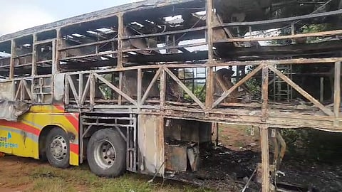 An external side-view photo of a bus that has been completely gutted by fire. The majority of the bus's body is charred and the windows are missing, revealing the skeletal metal framework of the upper and lower decks. The front of the bus is not visible, but a portion of the yellow and red painted body near the back wheels is partially intact, showing a few wheels. The bus is resting on the side of a road with green grass and dirt visible in the foreground. The background is somewhat blurred with foliage and trees.