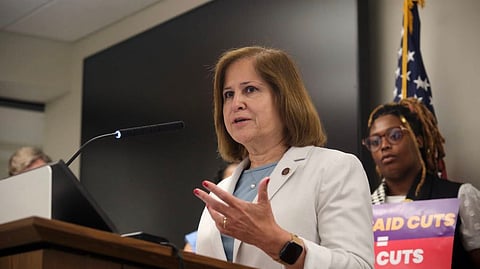 Virginia State Senator Ghazala Hashmi, a woman with shoulder-length brown hair, speaks firmly at a wooden podium, wearing a light-colored blazer. A person stands behind her on the right holding a sign that reads "AID CUTS = CUTS."