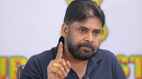 A man, identified as Indian actor and politician Pawan Kalyan, is seated at a table during a press conference.

He has a beard, is wearing a dark blue collared shirt, and is raising his right index finger as he speaks. He is looking off to his right with a serious expression. In the foreground, there are numerous microphones from different media outlets clustered on the table. A portion of the National Emblem of India is visible on the wall behind him.