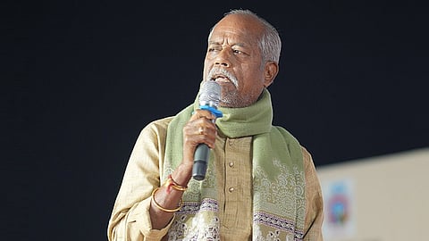An older man with a grey beard and mustache is speaking into a microphone. He is wearing a light brown or beige kurta and a light green scarf with a patterned border around his neck. He has a gold ring on his right hand, which is holding the microphone, and a few threads or bracelets on his wrist. The background is dark and out of focus.