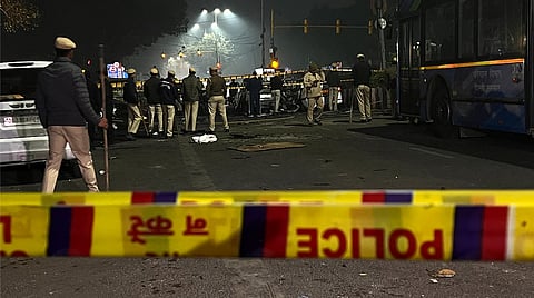 Delhi Police personnel stand near the site of a car explosion close to Red Fort Metro Station, with the area cordoned off by yellow police tape on a dimly lit night.