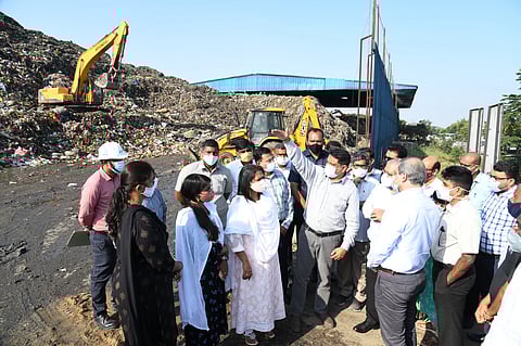 A group of officials, many wearing face masks, are gathered at a large dump site, likely the Jawaharnagar Dumpyard. In the background, there are enormous piles of waste being worked on by heavy machinery, including a yellow excavator on the left and a backhoe loader in the center. The people are engaged in a discussion, with one man in the center pointing towards the piles of garbage. The sky is bright and clear.