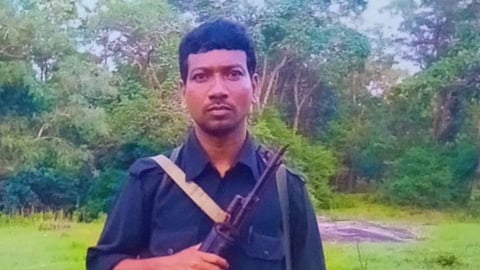 A medium-shot photograph of a man standing outdoors in a grassy, forested area. He is wearing dark blue or black clothing, appearing to be a button-down shirt and trousers. He is holding an assault rifle, likely an AK-47 variant, diagonally across his chest with a strap over his shoulder. He has an ammunition belt around his waist and is wearing sandals on his feet. The background consists of green grass and dense forest foliage with some rock formations visible in the distance.