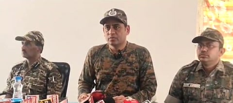 A photograph of three men in camouflage-patterned police or paramilitary uniforms, sitting at a table with microphones in front of them, seemingly during a press conference. The central figure, who appears to be speaking, is wearing a cap. The man on the right has "AMIT BADURIA" visible on his uniform name tag. The man on the left is partially visible.