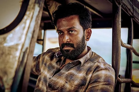 Prithviraj Sukumaran as Double Mohanan, sitting inside a vehicle, wearing a checked shirt, with a rugged beard, darkened skin tone, and an intense, brooding expression.