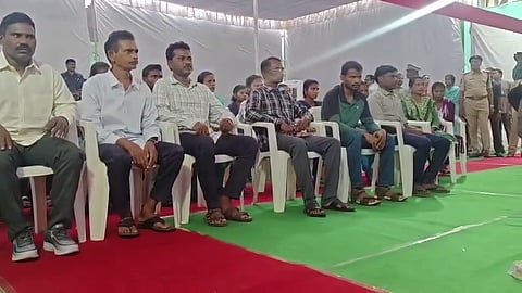 A group of people, mostly men, are seated in white plastic chairs under a tent structure. They are sitting in a row facing forward. Many of the men are wearing light-colored button-up shirts and dark trousers. There are other people standing behind them, including what appears to be a police officer on the right side of the frame. The floor is covered with a green and red carpet.