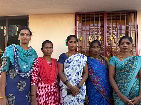 An image of five women standing side-by-side outdoors in front of a building. The women, who appear to be Indian, are wearing colorful saris and traditional attire. Their expressions are serious or solemn. The women are described in the file name as "Families of the fishermen."