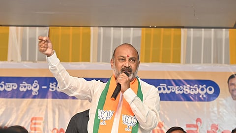 A horizontal, close-up photograph shows an Indian male politician, Bandi Sanjay Kumar, addressing a rally or crowd. He is middle-aged, with a graying beard and mustache, dressed in a white kurta and draped with an orange and green kanduva (shawl or stole), which features the lotus symbol of the Bharatiya Janata Party (BJP). He is holding a microphone near his mouth with his right hand and raising his left arm and fist high in a gesture of address or emphasis. He has a serious expression. Behind him is a large banner, partially visible, with text in the Telugu language and the BJP's yellow, white, and blue colors. In the foreground, below him, the tops of the heads of several attendees are visible, including one man looking up intensely on the bottom right.