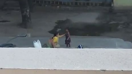 A low-resolution, slightly blurry image taken from an elevated perspective, looking over a pale tan wall or ledge. In the distance, below the ledge, two small figures, likely children, are visible outdoors. One figure, wearing yellow, appears to be holding hands with or tending to the other figure, who is wearing a dark, possibly maroon, outfit. Behind them is a dirt ground area, and further back, there are trees and a blue and grey building structure. The image is cropped significantly by the foreground wall.