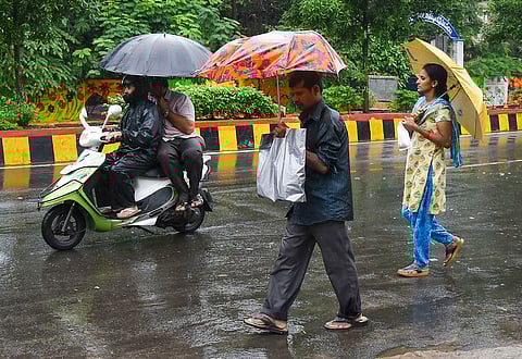 A street scene in Visakhapatnam, India, showing people navigating heavy rain. On the left, two men on a white and neon-green scooter are fully covered in black rain gear and sheltered by a large black umbrella, driving on the wet road. In the center, a man walks with an orange and floral-patterned umbrella, carrying a large silver bag. On the right, a woman walks holding a yellow umbrella, wearing a yellow kurta with a blue pattern and blue jeans. The road is slick with water, and a painted curb is visible behind them, separating the road from a grassy, tree-lined area.