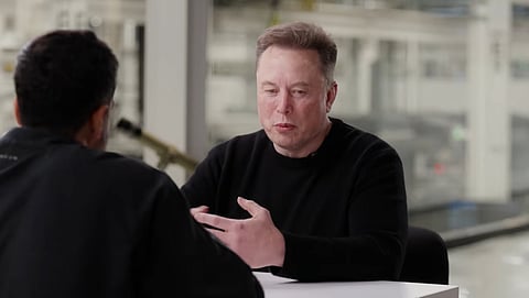 Elon Musk, dressed in a black full-sleeved shirt, sits at a table. Podcaster and businessman Nikhil Kamath is also in the frame, but the camera is behind him. The interior of the building can be seen in the background blurrily. 