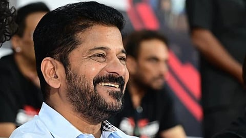 A vertical close-up photograph shows a smiling man, presumably Revanth Reddy, seated in an indoor setting. He has a dark beard, mustache, and short dark hair. He is wearing a light blue, long-sleeved collared shirt with a subtle, dark plaid pattern visible on the inside of the collar. He is looking off to the side, his hands clasped in front of him. The background is dark and blurred, with partially visible people and some red and black elements suggesting he is at an event.