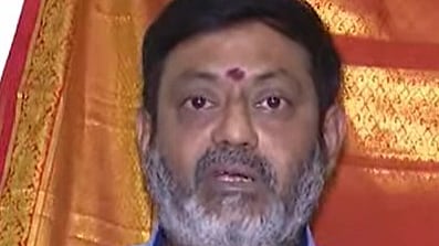 A middle-aged man with a short, salt-and-pepper beard is looking directly at the camera. He is wearing a light blue collared shirt with a small emblem on the chest. He has a red tilak mark on his forehead, and a backdrop of orange and red fabric is visible behind him.