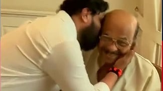 Raman Pillai is seated in a chair. Dileep is standing next to him and bends down to hug Raman Pillai.