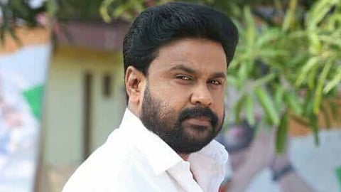 A close-up photograph of the Malayalam actor Dileep (Gopalakrishnan Padmanabhan Pillai). He has a dark beard and mustache, dark hair, and is wearing a white collared shirt. He is looking directly at the camera with a serious expression. The background is slightly blurred and appears to be outdoors.