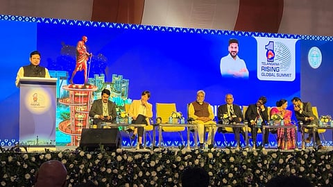 
A low-angle photo captures a man speaking at a podium on a large stage, flanked by several dignitaries seated in a row.

The speaker, wearing a dark jacket over a white shirt, is to the left, standing behind a podium with the 'Telangana Rising Global Summit' logo. The backdrop is a large LED screen. On the screen, a graphic of a standing Mahatma Gandhi statue is visible to the speaker's left, and to the speaker's right, a portrait of a man (likely the Chief Minister or another prominent leader) is displayed next to the "Telangana Rising Global Summit" logo.

Seven people are seated on yellow chairs along the stage, facing the audience. They include men in both traditional Indian wear and suits, and one woman in a pink floral saree. The stage is decorated with white and purple floral arrangements along the front edge, and a large crowd is partially visible in the foreground.