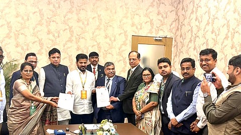 A group of people, primarily men in business attire or traditional Indian wear, are gathered around a large boardroom table. In the center, a woman in a saree is handing a document to a younger man in a white and gold outfit (possibly a kurta) who is holding another document. Several other people are surrounding them, watching the exchange and smiling. Two men on the far right are taking photos with mobile phones. The setting is a formal meeting room with light-colored, patterned wallpaper. The image appears to capture an agreement or partnership being formally signed or exchanged.
