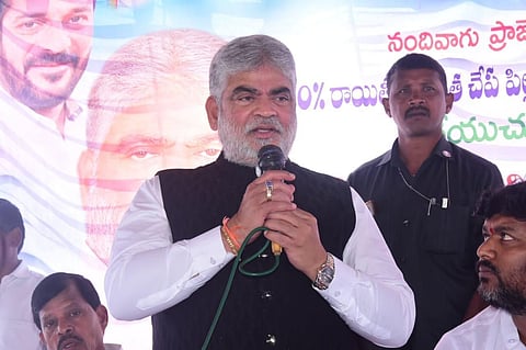 A middle-aged Indian man with a white beard and grey hair, wearing a white shirt and a black Nehru vest, speaks into a microphone held with both hands. He is at an outdoor event with a banner in the background featuring Telugu script and a portrait of another man. Several other men are visible in the background and foreground, including one in a security uniform.
