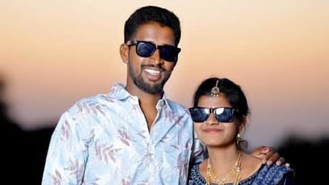 A medium shot of a young man and woman standing close together outdoors during sunset.

The man, on the left, has a dark complexion, a short beard, and is smiling. He is wearing a light blue button-down shirt with a subtle leaf pattern and dark sunglasses.

The woman, on the right, is wearing a traditional dark blue and silver lehenga with gold jewelry, including two necklaces, a waist belt, and a maang tikka on her forehead. She is also wearing dark sunglasses and has her arm resting behind the man.

The background is a soft, blurred golden-orange sky, suggesting dusk.