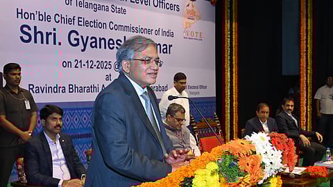 A medium shot of Shri Gyanesh Kumar, the Chief Election Commissioner of India, speaking at a podium during an event in Hyderabad on December 21, 2025.

He is a middle-aged man with graying hair and glasses, wearing a dark grey suit and a patterned blue tie. He stands behind a wooden podium heavily decorated with vibrant orange, yellow, and white marigold garlands.

In the background, a large white banner displays his name and title, along with the text "Ravindra Bharathi, Hyderabad." Several other officials in formal attire are seated on the stage behind him, and the setting is formal, featuring traditional floral decorations and green carpeting.