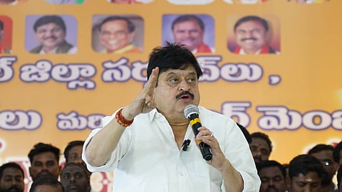 An image showing N. Ramchander Rao, a senior leader of the Bharatiya Janata Party (BJP) in Telangana, addressing a gathering. He is wearing a white shirt and holding a microphone in his left hand while gesturing with his right hand during a speech.

In the background, there is a large orange banner featuring Telugu text and portraits of other political figures. Several people are seated behind him, listening to the address. The setting appears to be a formal political event or meeting.